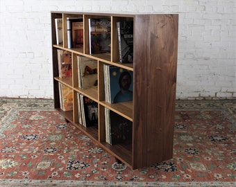 May include: A walnut wood bookshelf with 12 compartments, filled with books and vinyl records. The bookshelf is standing on a patterned rug.