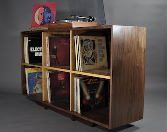 May include: A walnut wood record player stand with six shelves, each filled with vinyl records. The stand has a light wood top and a turntable on top. The records include titles like "Electra Music" and "Pierre Moerlen Returns to the Village Gate".