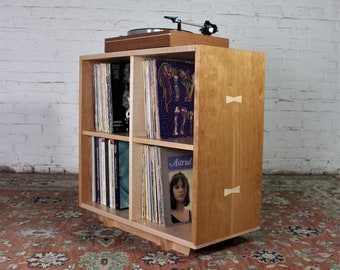 Puede incluir: Un soporte de tocadiscos de madera clara con cuatro estantes para guardar discos de vinilo. El soporte tiene un acabado de madera natural y presenta dos pequeños detalles de madera en la parte delantera. El soporte está sobre una alfombra estampada.