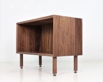 May include: A wooden shelf unit with a single open shelf. The shelf is made of walnut wood and has a natural finish. The shelf is supported by four legs, each with a brass foot.