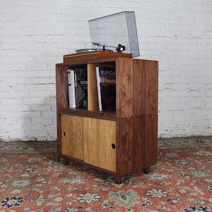 May include: A two-tiered walnut wood record player stand with sliding doors. The top shelf has a record player and a stack of books. The bottom shelf has sliding doors.
