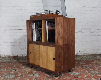 May include: A two-tiered walnut wood record player stand with sliding doors. The top shelf has a record player and a stack of books. The bottom shelf has sliding doors.