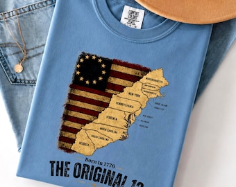 Könnte beinhalten: Ein hellblaues T-Shirt mit Rundhalsausschnitt und einer Grafik der ursprünglichen dreizehn amerikanischen Kolonien auf einer Flagge im Used-Look. Der Text lautet Born In 1776 THE ORIGINAL 13, inklusive Beschriftungen.