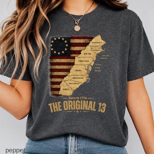 May include: A charcoal grey t-shirt featuring a graphic of the American flag with a circle of thirteen stars. Overlaid is a map of the original thirteen colonies with state names listed. Text reads: Born In 1776 THE ORIGINAL 13.