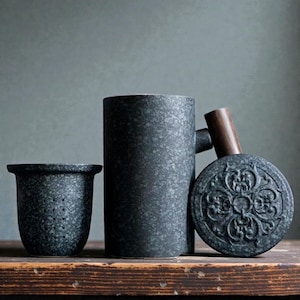 May include: A dark gray tea set with a speckled texture. The set includes a small strainer, a tall cylindrical mug, and a round lid with a wooden handle. The lid has an embossed floral design. The items are arranged on a wooden surface.