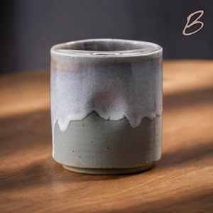 May include: A handcrafted ceramic tumbler with a unique glaze. The top half is a light gray with a white, dripped effect, transitioning to a darker gray base. The tumbler sits on a wooden surface, showcasing its handmade quality.