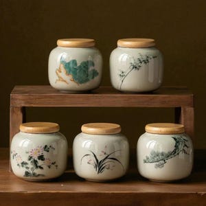 Pode incluir: Cinco potes de cerâmica com tampas de madeira são exibidos em uma prateleira de madeira. Cada pote é decorado com um design botânico pintado à mão diferente em tons de verde, rosa e preto. Os potes têm um acabamento brilhante.