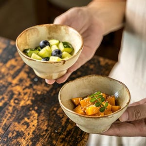 May include: Two small, round, off-white ceramic bowls with a rustic, speckled finish and uneven rims. One bowl contains diced cucumbers and blueberries, while the other holds diced orange squash topped with a small green sprig of herbs.