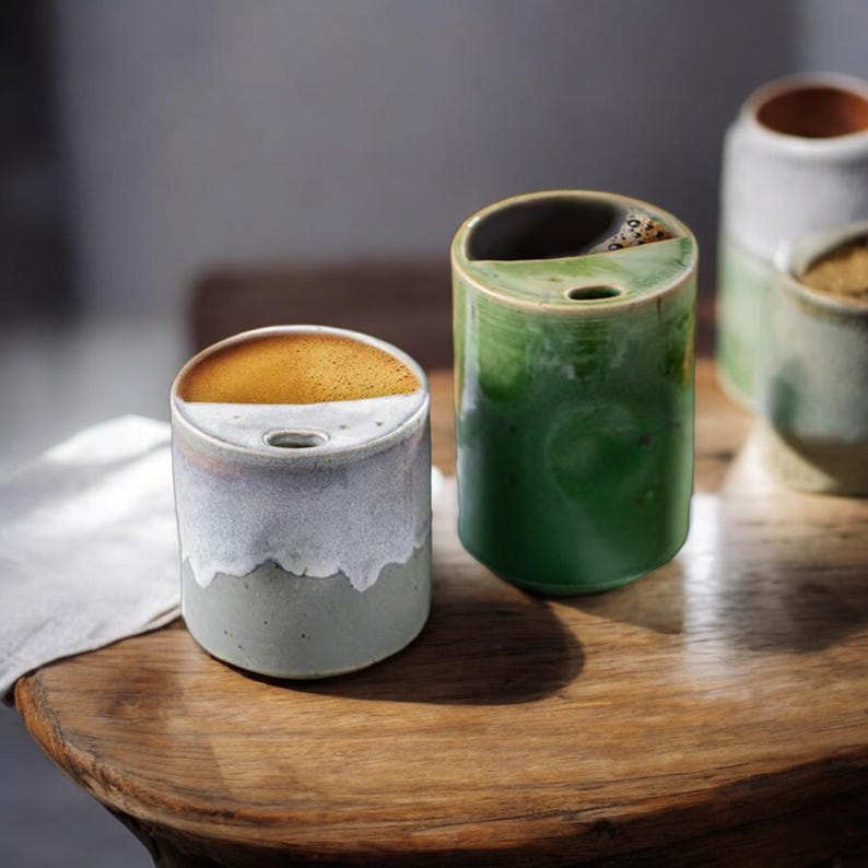 May include: A collection of ceramic mugs in various colors, including a two-toned gray and white mug with a brown interior and a green mug. The mugs are cylindrical with a small hole on the top. The mugs are on a wooden surface.