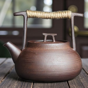 May include: A brown ceramic teapot with a rope-wrapped handle. The teapot has a spout and a small lid with a handle. The teapot is sitting on a wooden surface.