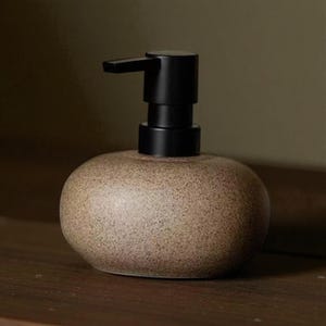 Könnte beinhalten: Ein gesprenkelter, brauner Keramik-Seifenspender mit schwarzer Pumpe. Der Spender hat eine abgerundete, bauchige Form und steht auf einer Holzoberfläche. Die Pumpe befindet sich oben auf dem Spender.