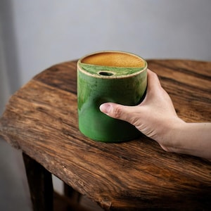 May include: A green ceramic mug with a unique design, held by a hand. The mug has a cylindrical shape with a curved top and a small hole. The interior is filled with a tan liquid. The mug is on a wooden table.