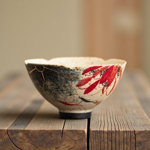May include: A decorative ceramic bowl. The bowl has a cream interior and a dark blue and gold exterior with red leaf patterns. The bowl is placed on a wooden surface, highlighting its intricate design and craftsmanship. The bowl's diameter is approximately 15 cm.