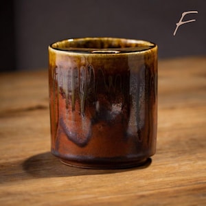 May include: A cylindrical ceramic mug with a rich, earthy color palette. The mug features a dark brown base that transitions to reddish-brown and then to a glossy, dark brown rim. The glaze has a dripped effect, adding visual interest. The mug sits on a wooden surface.