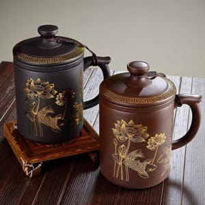 May include: Two ceramic tea mugs with lids. One is black, the other is brown. Both have gold lotus and koi fish designs. Each mug has a handle and a decorative band around the lid. The mugs sit on a wooden tray.