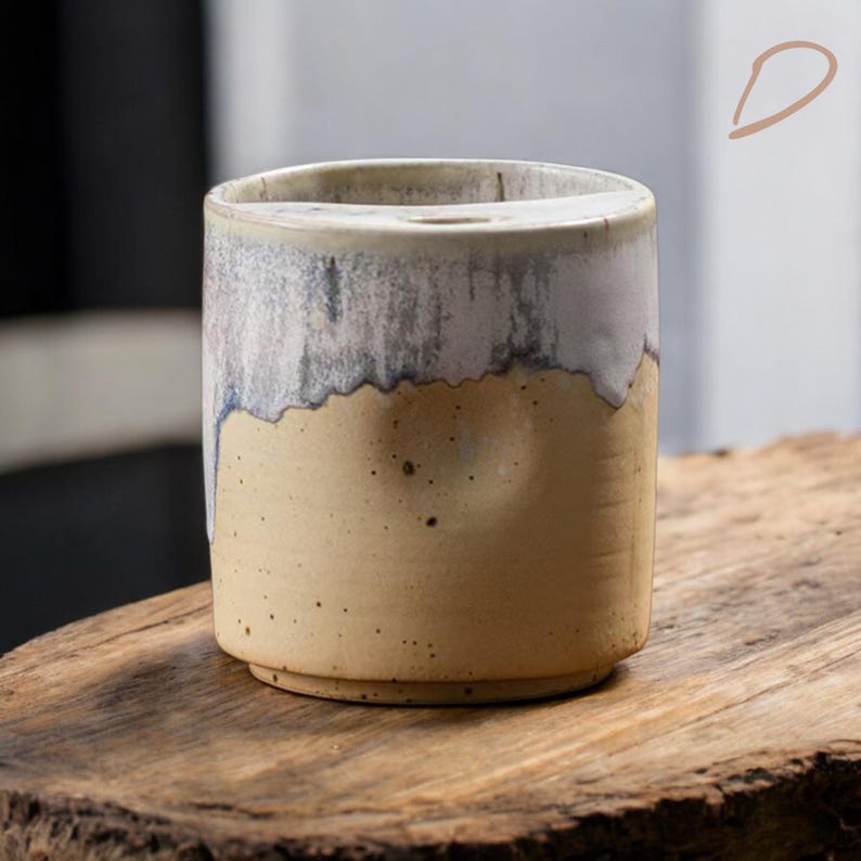 May include: A cylindrical ceramic vase with a two-tone design. The top half is a light gray with a textured, dripped effect, while the bottom half is a speckled tan. The vase sits on a wooden surface.