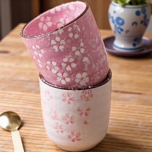 Puede incluir: Dos tazas de cerámica apiladas. La taza superior es rosa con diseños florales blancos, y la inferior es blanca con diseños florales rosas. Ambas tazas tienen un borde marrón.
