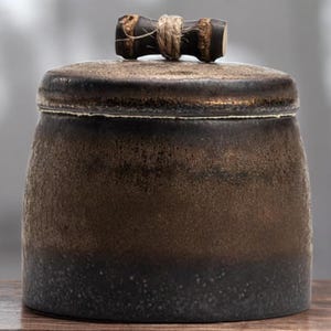 Puede incluir: Un tarro de cerámica con tapa, con un degradado marrón oscuro y negro. El tarro tiene una superficie texturizada y un asa de estilo bambú atada con cuerda. El diseño sugiere una estética rústica o artesanal, adecuada para almacenar.