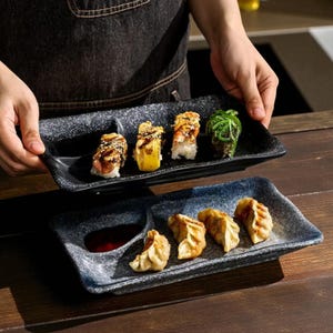 Puede incluir: Dos platos rectangulares de sushi con un diseño moteado en blanco y negro. El plato superior contiene rollos de sushi con varios ingredientes. El plato inferior tiene empanadillas y un cuenco pequeño de salsa roja.
