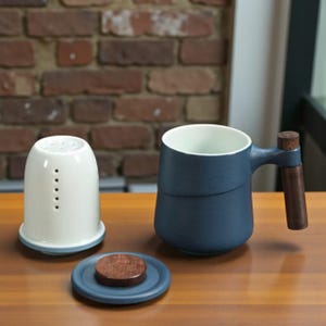May include: A modern tea set featuring a navy blue mug with a wooden handle, a white tea infuser, and a small blue saucer with a wooden lid. The mug has a white interior and a unique design. The set is on a wooden surface.