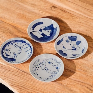 May include: Four small, white ceramic plates with blue cat illustrations. Each plate features a different sleeping cat design. The plates are arranged on a wooden surface, showcasing the detailed artwork and playful theme.