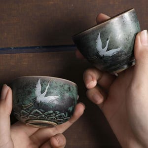 Puede incluir: Dos tazas de té de cerámica con un esmalte verde azulado y marrón. Cada taza presenta un diseño de grulla blanca. Las tazas se sostienen en las manos, mostrando la obra de arte detallada y la forma de las tazas. Las tazas tienen una apariencia artística única.