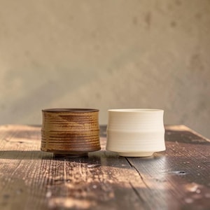 May include: Two cylindrical ceramic cups sit side by side. The cup on the left features a textured, dark brown finish with horizontal ridges. The cup on the right has a smooth, matte off-white finish with a subtle horizontal band near the base.