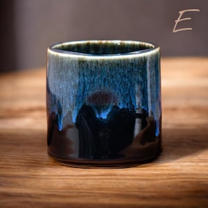May include: A ceramic tumbler with a unique glaze. The cup features a dark base that transitions to shades of blue and teal, with a glossy finish. The rim has a dark brown color. The tumbler is cylindrical and sits on a wooden surface.