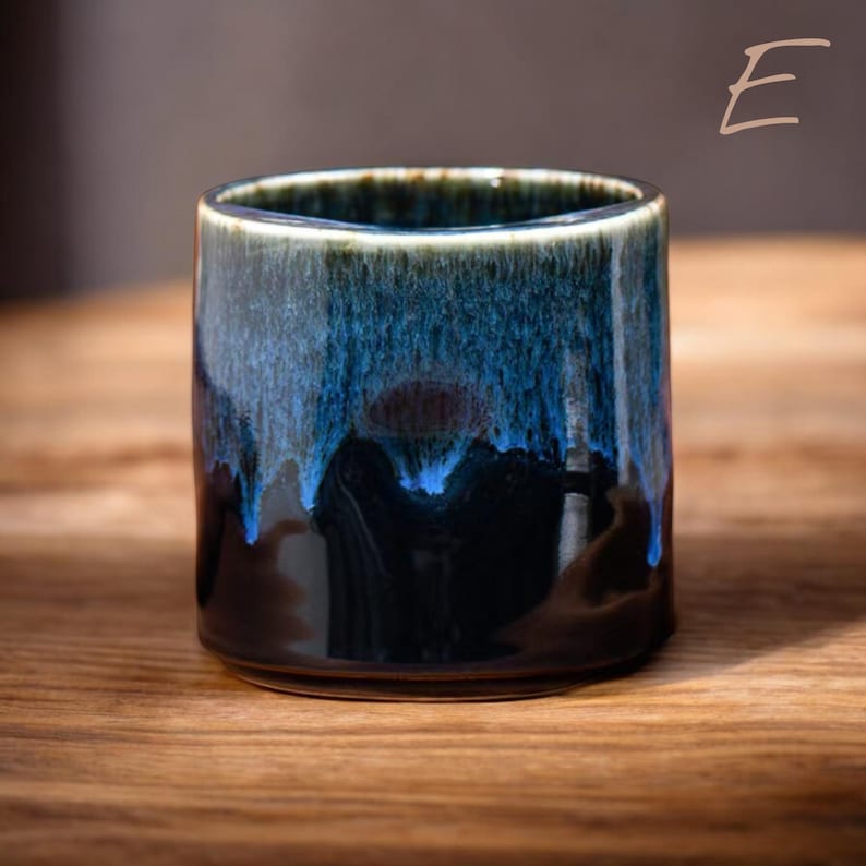 May include: A ceramic tumbler with a unique glaze. The cup features a dark base that transitions to shades of blue and teal, with a glossy finish. The rim has a dark brown color. The tumbler is cylindrical and sits on a wooden surface.