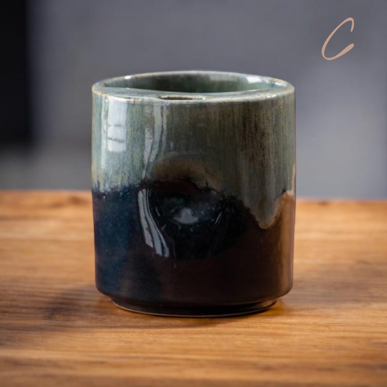 May include: A cylindrical ceramic container with a dark blue and green glaze. The top portion is green, transitioning to dark blue at the base. The container sits on a wooden surface.