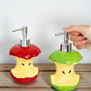 May include: Two ceramic soap dispensers shaped like apple cores. One is red, the other green, with yellow interiors and silver pump tops. The dispensers are on a wooden surface.
