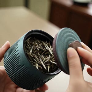 Pode incluir: Um recipiente de chá de cerâmica verde-azulado com um exterior canelado e uma tampa a condizer. O recipiente está aberto, revelando folhas de chá secas no interior. A tampa está a ser levantada, mostrando uma pega de madeira castanha.
