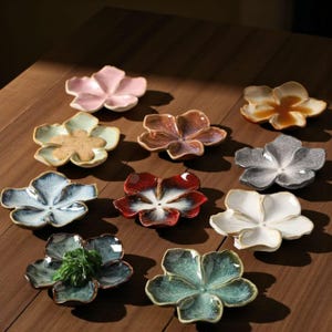 Pode incluir: Uma coleção de pratos de cerâmica em forma de flor em várias cores, incluindo rosa, azul, castanho e branco. Os pratos estão dispostos sobre uma superfície de madeira, mostrando o seu design floral único e acabamento brilhante. Alguns pratos contêm pequenas plantas verdes.