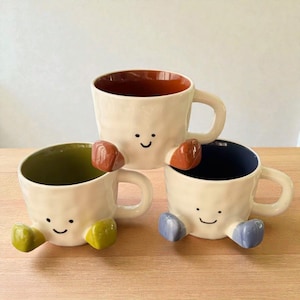 May include: Three quirky ceramic mugs with smiling faces and colourful accents. Each mug has a different interior colour: brown, green, and blue. The mugs have small, rounded appendages in various colours, adding to their playful design.