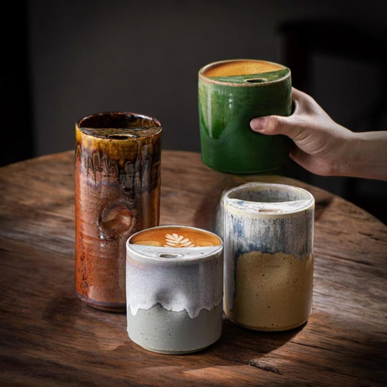 May include: Four ceramic coffee mugs with a unique design. The mugs are cylindrical with a small opening at the top. The mugs have a variety of colors, including brown, green, gray, and blue. One mug is being held by a person.