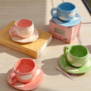 Könnte beinhalten: Vier Keramik-Tassen und Untertassen in Pastellfarben. Eine rosa Tasse hat ein Karomuster, während andere Wolkenmotive aufweisen. Die Tassen stehen auf einer Holzoberfläche, mit einem Buch und einer dekorativen Box in der Nähe.