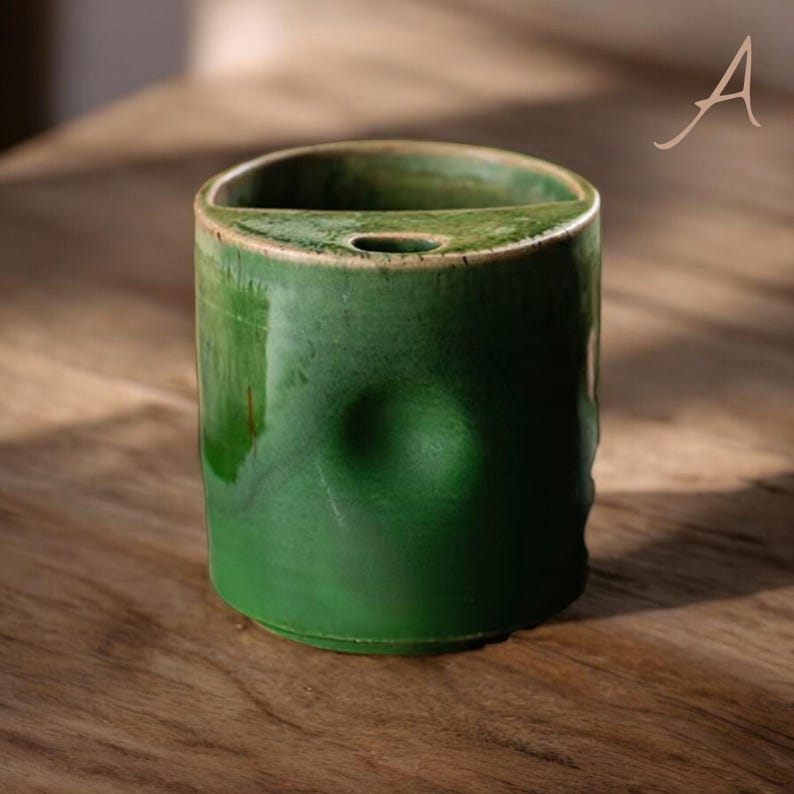 May include: A cylindrical, handmade ceramic container with a glossy, emerald green glaze. The top has a curved opening and a small hole. The container has a unique, dimpled design on the front. The piece is set on a wooden surface.