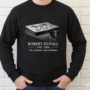 May include: Black sweatshirt featuring a graphic of a book, eyeglasses, and a glass of whiskey. The text reads "ROBERT DUVALL 1931-2026" and "I'm a lawyer, not a hitman."