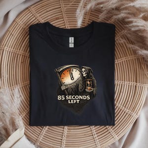 May include: Black t-shirt featuring a graphic of the Grim Reaper holding a scythe and hourglass, with a clock face showing the time. The text "85 SECONDS LEFT" is below the graphic. The shirt is folded on a woven surface.