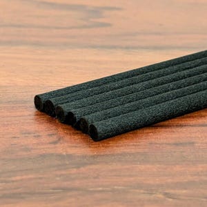 May include: A close-up of a bundle of black diffuser sticks, arranged diagonally across a wooden surface. The sticks are cylindrical and have a textured, porous appearance. The background is a warm, medium-toned wood.