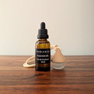 Pode incluir: Um frasco de vidro âmbar com tampa conta-gotas preta e rótulo preto com o texto RADIANCE Essential oils Car air freshener 30ml. Ao lado, um pequeno difusor de vidro fosco com tampa de madeira clara e um cordão bege para pendurar.