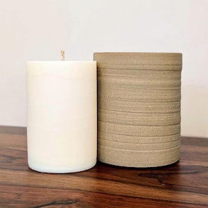 May include: A cylindrical white wax candle with a single centred wick positioned next to a cylindrical beige ceramic vessel featuring a textured exterior with horizontal grooves and a matte finish, both standing upright on a flat surface.