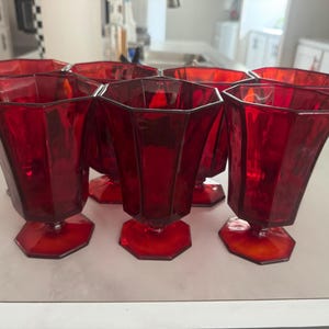 Op de afbeelding: Een set van zes robijnrode glazen bekers met een gefacetteerd, meerzijdig ontwerp, een zeshoekige voet en een korte steel. Elk drinkglas heeft een diepe, doorschijnende karmozijnrode tint en een gestructureerd, geometrisch silhouet met verticale panelen.