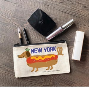 May include: A canvas cosmetic bag with a black zipper, featuring a cartoon dachshund designed as a hot dog with the text "NEW YORK". The bag also has a black compact, mascara, a perfume bottle, and two pens.