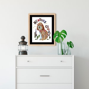 Op de afbeelding: Een ingelijst afdruk van een hond met een bril en een rode bandana met de woorden "NEW YORK CAVAPOO" in kleurrijke letters. De hond staat voor een stadsbeeld.