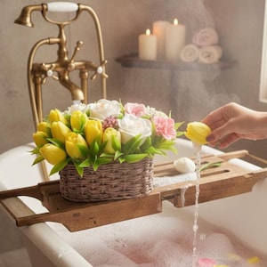 May include: A woven basket of yellow tulips, white and pink roses, and green foliage rests on a wooden bath tray. A hand holds a yellow tulip over the water, creating a stream. The bathtub has a vintage brass tap.