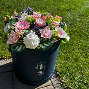 May include: A vibrant floral arrangement in a navy blue round box. The bouquet features a mix of pink, white, and purple flowers with green foliage. The box has a logo that reads "The Flowers". The arrangement is set against a backdrop of green grass.