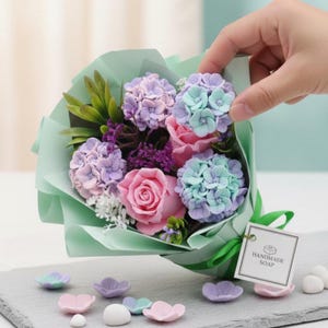May include: A bouquet of handmade soap flowers in shades of pink, purple, and blue, wrapped in light green paper. The bouquet includes roses, hydrangeas, and other floral elements. A tag reads "HANDMADE SOAP".