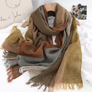 Puede incluir: Bufanda multicolor en tonos marrones, grises y beige, con bordes deshilachados. La bufanda está drapeada sobre una percha de madera, mostrando su textura suave y variaciones de color. La bufanda parece estar hecha de un material ligero.