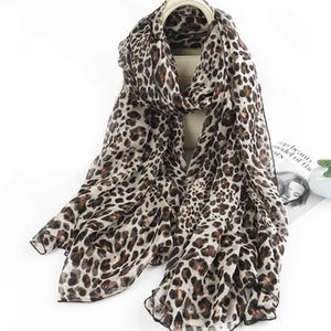 Könnte beinhalten: Leichter Schal mit Leopardenmuster in Braun-, Beige- und Schwarztönen. Der Schal ist über einer neutralfarbenen Tasche drapiert, wodurch der fließende Stoff und das Design hervorgehoben werden. Die Kanten des Schals sind mit dunklem Besatz versehen.